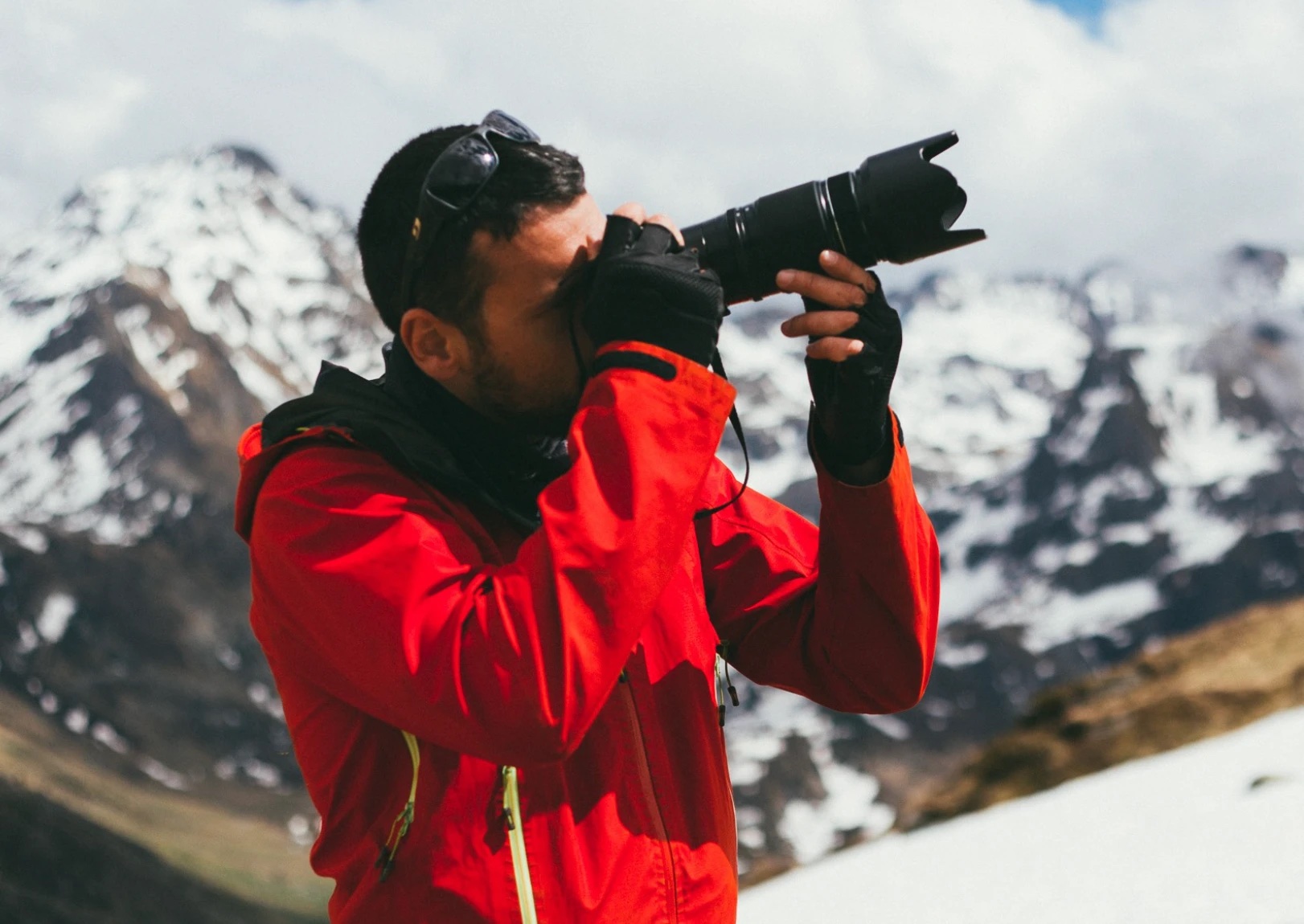 Photographe en veste rouge, l'œil au viseur, devant des sommets enneigés